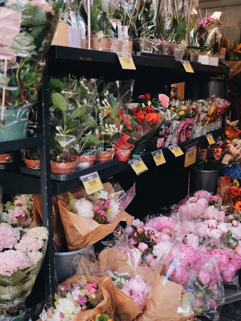 A floral display inside a flower shop showcases a variety of fresh flowers arranged on black metal shelves. The top shelf features potted plants with green leaves and flowering arrangements wrapped in