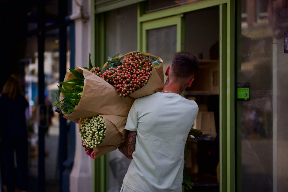 A person with short dark hair, wearing a plain white t-shirt, is seen from behind carrying a large bouquet of fresh flowers wrapped in brown paper. The bouquet includes clusters of pink berries, white