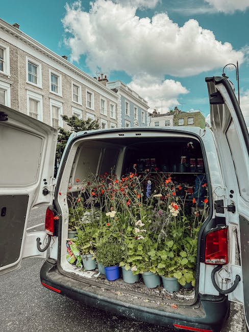The back of a delivery van with the rear doors open, revealing an assortment of potted plants and flower stems with red, pink, and green foliage, ready for floral arrangements or flower delivery. The 