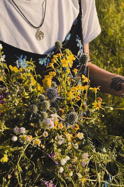 A close-up of a person holding a vibrant mixed bouquet of fresh wildflowers, including yellow daisies, purple thistles, and small white blossoms, arranged loosely with visible green foliage. The indiv