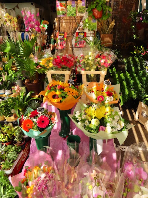 A vibrant floral display featuring various fresh flower bouquets arranged on a pink cloth-covered table inside a flower shop. The bouquets include bright red roses, yellow sunflowers, pink gerbera dai