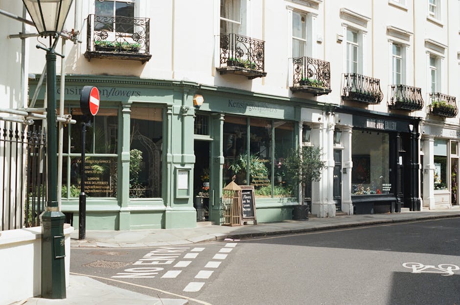 A charming florist shop with a light green exterior situated on a street corner, featuring large glass windows displaying various floral arrangements. The shop's sign includes the words 'Kensington Fl