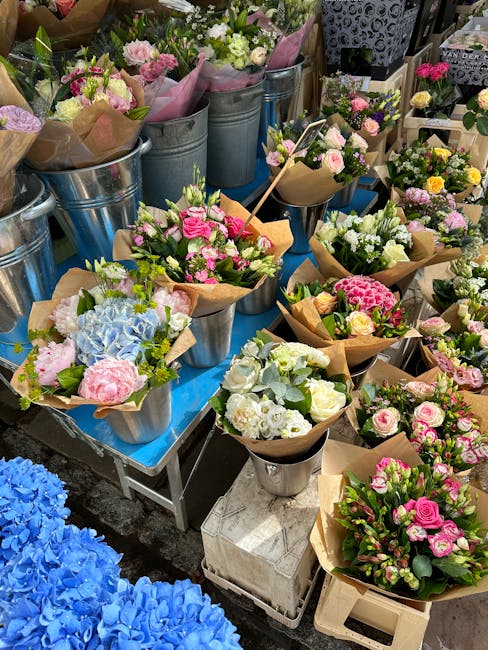 An array of freshly arranged flower bouquets displayed on a blue table at a flower market. The arrangements feature a variety of blossoms including pink roses, white hydrangeas, and pastel carnations,