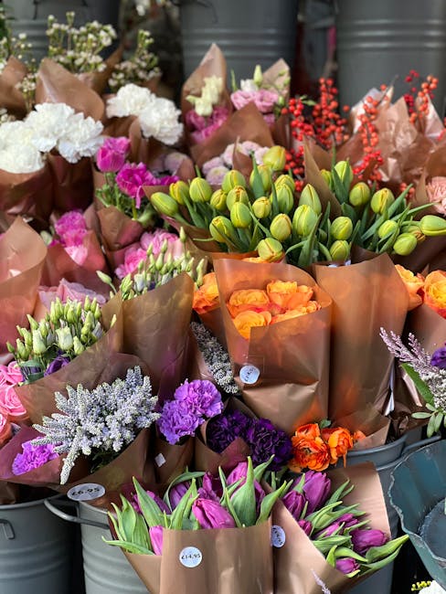 A vibrant display of freshly cut flowers, including pink tulips, purple lisianthus, orange roses, white astilbe, and closed yellow tulip buds, all arranged in brown paper wrapping. The bouquets are re