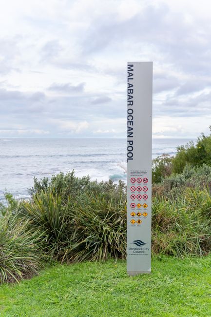 A tall, vertical sign reading 'Malabar Ocean Pool' stands on a grassy area near the coastline, with lush green shrubs and plants in the foreground. Behind the sign, the ocean stretches out to the hori