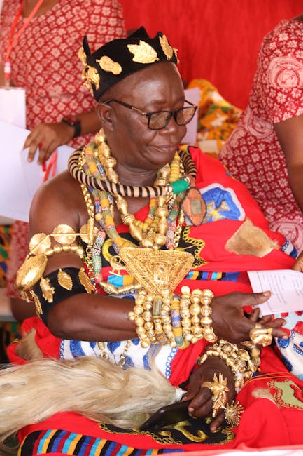 A woman dressed in traditional Maasai attire, wearing vibrant red and blue shuka cloth, adorned with elaborate gold jewelry including necklaces, bracelets, and earrings, sitting indoors against a red